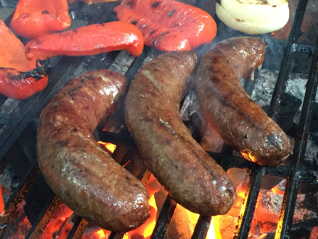 Photo of 3 sausage links, sliced red bell pepper and onion cooking on a barbeque grill