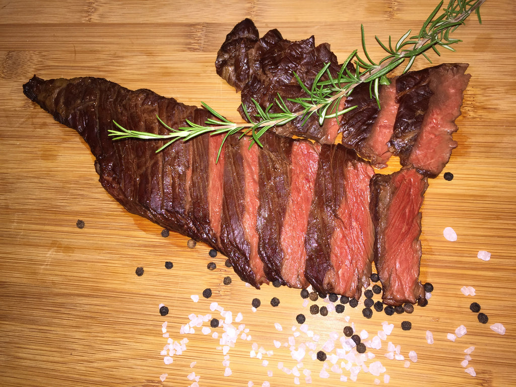 Photo of sliced rare flank  steak with pepper corns,  kosher salt and sprig of rosemary on cutting board