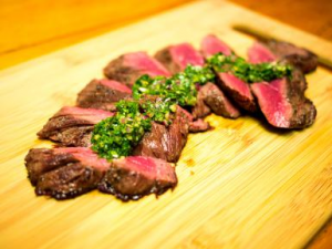 Photo of cooked rare hanger steak on a cutting board, sliced with chimichurri green sauce on top