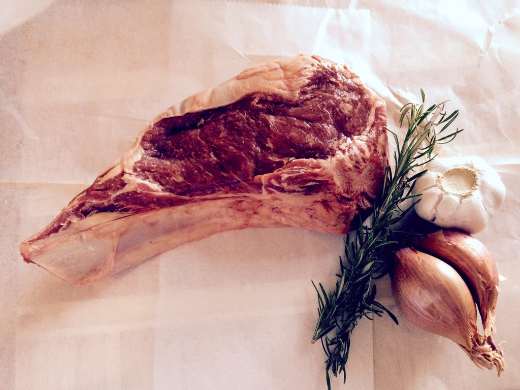 Photo of bone-in raw rib eye steak on white butcher paper with a sprig of rosemary, shallots and a bulb of garlic