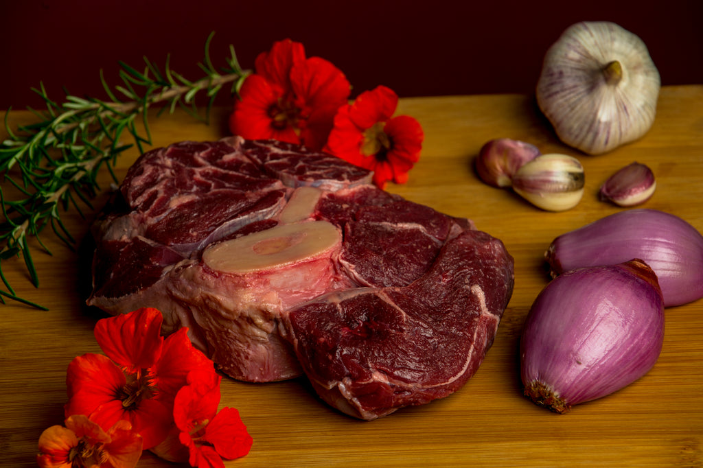 Photo of raw shank on a wood cutting board surrounded by shallots, garlic cloves and bulb , sprig of rosemary and red flowers