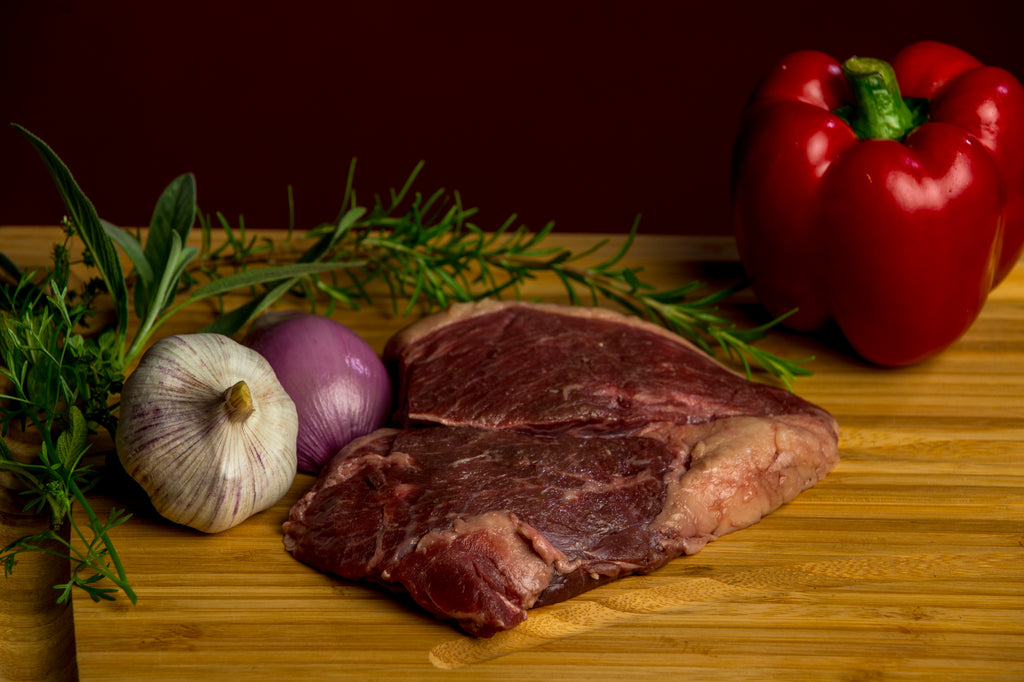Photo of a raw top sirloin steak with a whole red bell pepper, garlic bulb , shallot and sprig of rosemary on a wood board