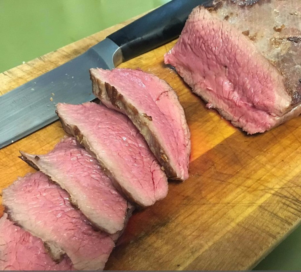 Photo of rare sliced tri tip roast on a wood cutting board with a black handled carving knife and green background