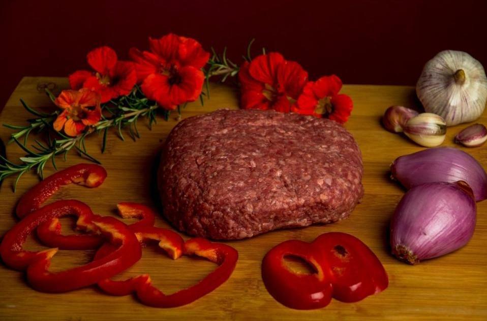Photo of raw hamburger patty on a wood board surrounded by red sliced bell peppers, shallots, garlic bulb and red flowers